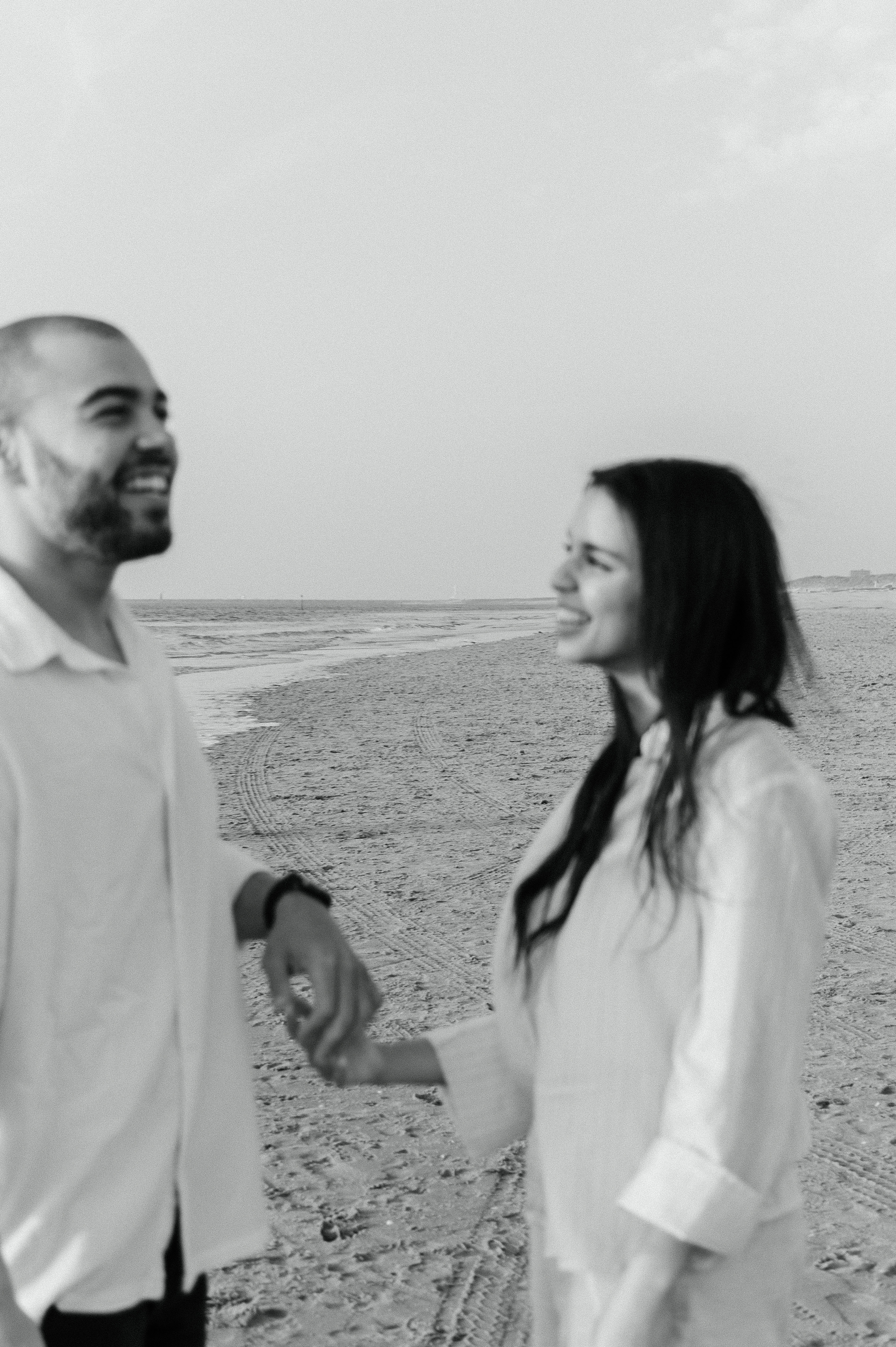 black and white photo of a couple standing on the beach holding hands with them out of focus and the background in focus.