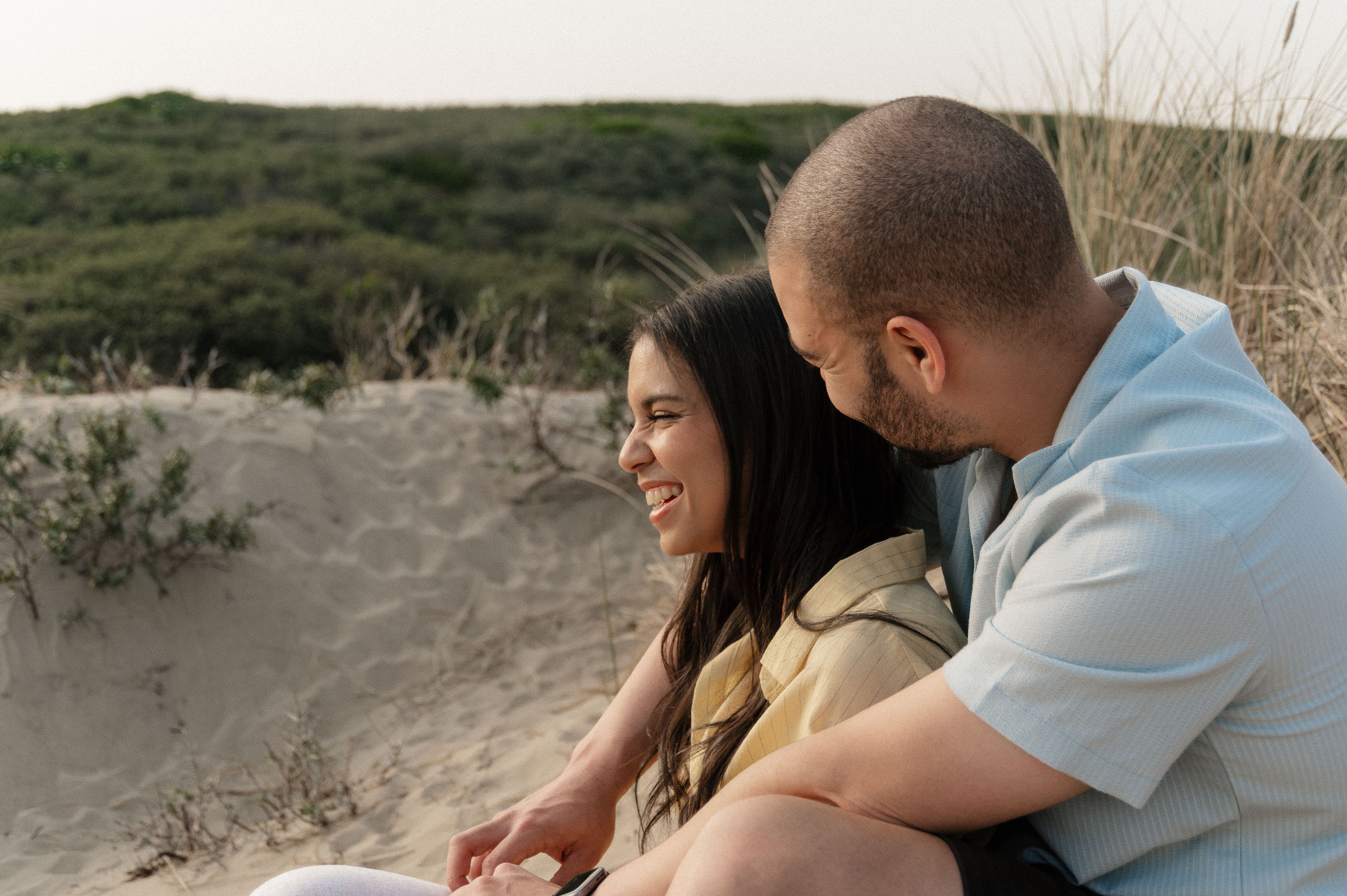 Close up shot of couples sitting on sand dune with girl sitting in between guys legs and both laughing.