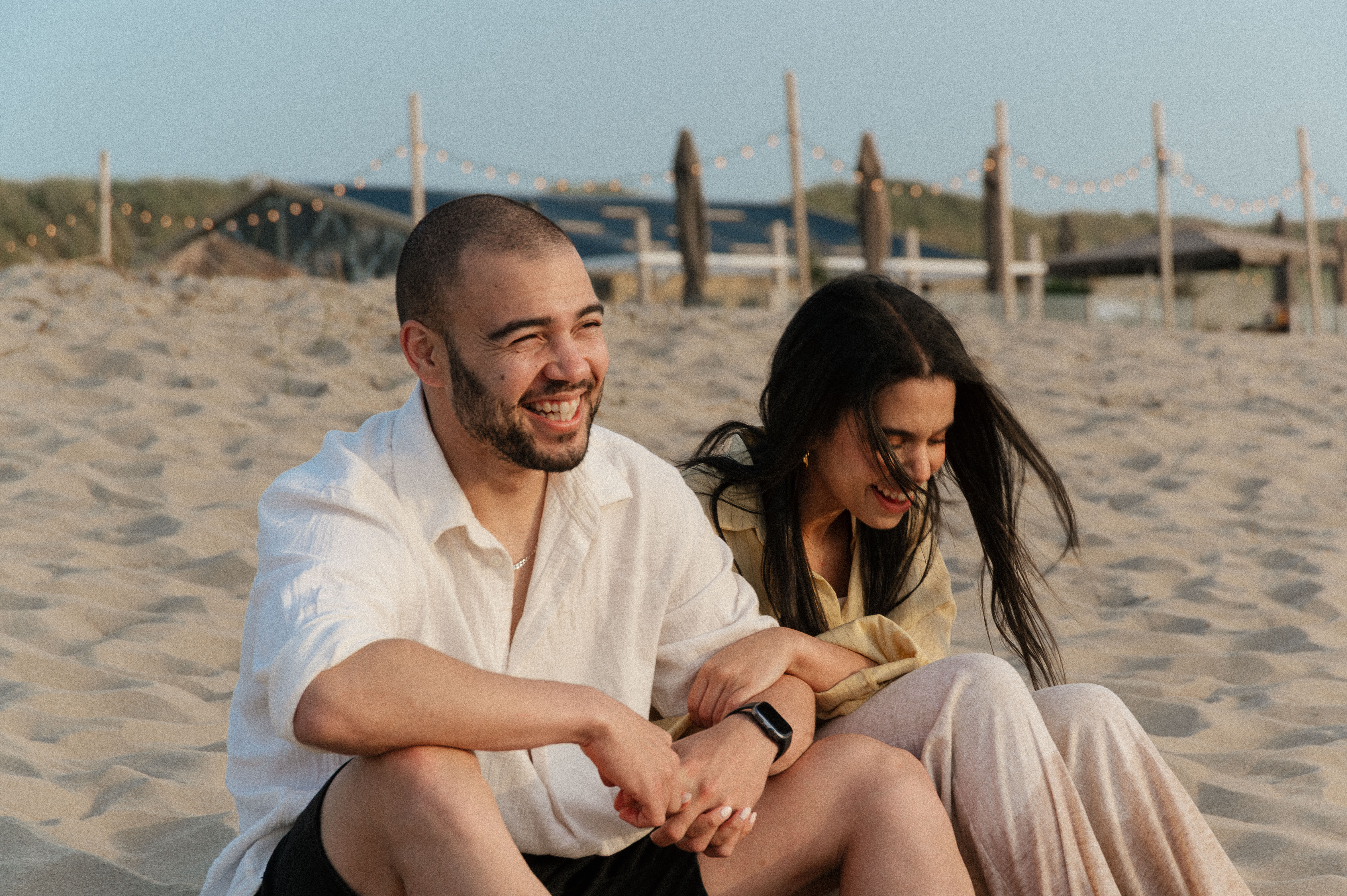 Couple sitting on beach next to each other with arms hooked in laughing together.