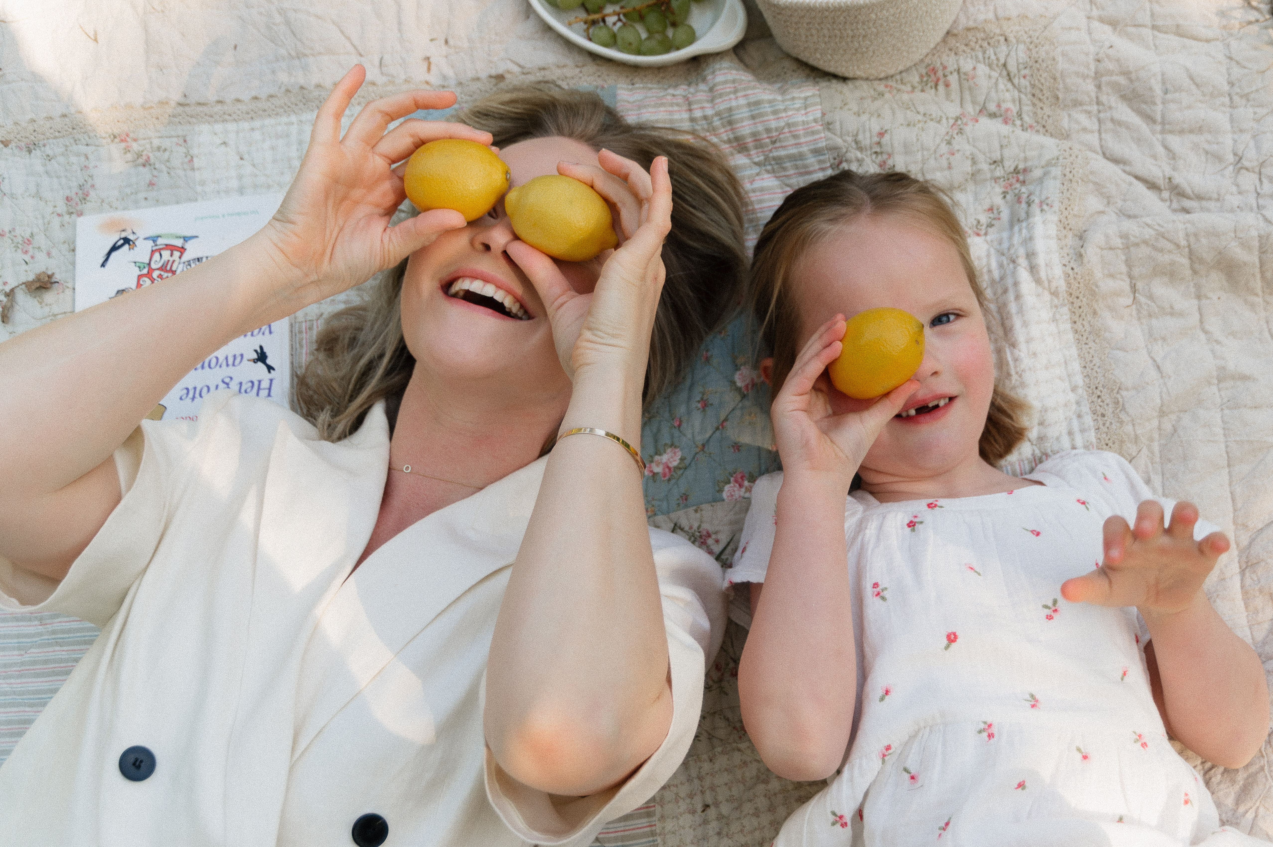 mother and daughter lying on their backs on a blanket holding lemons in front of their eyes.