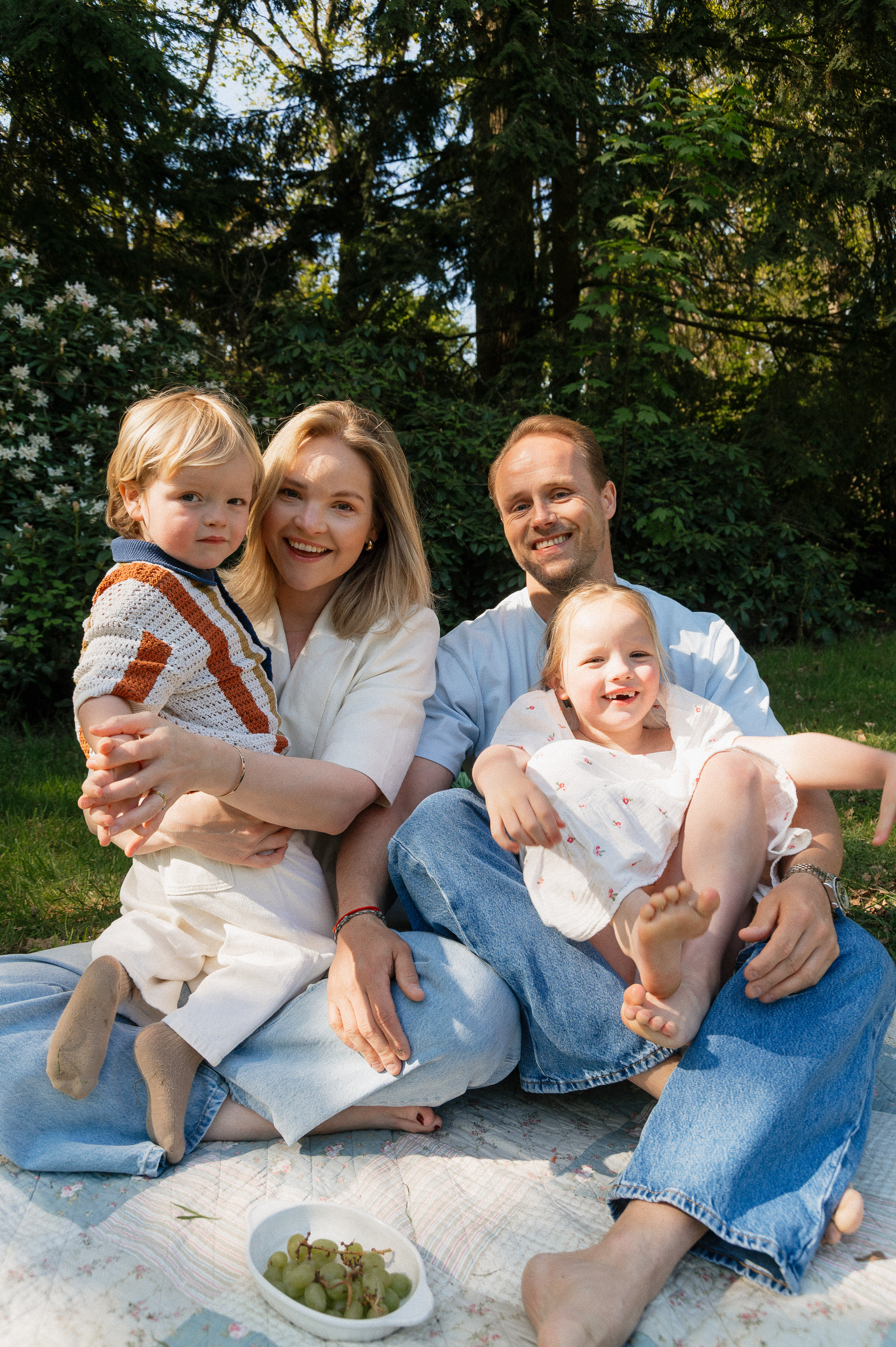 candid family portrait of mother, father, 6-year old daughter and 3 year old son sitting on blanket outside.