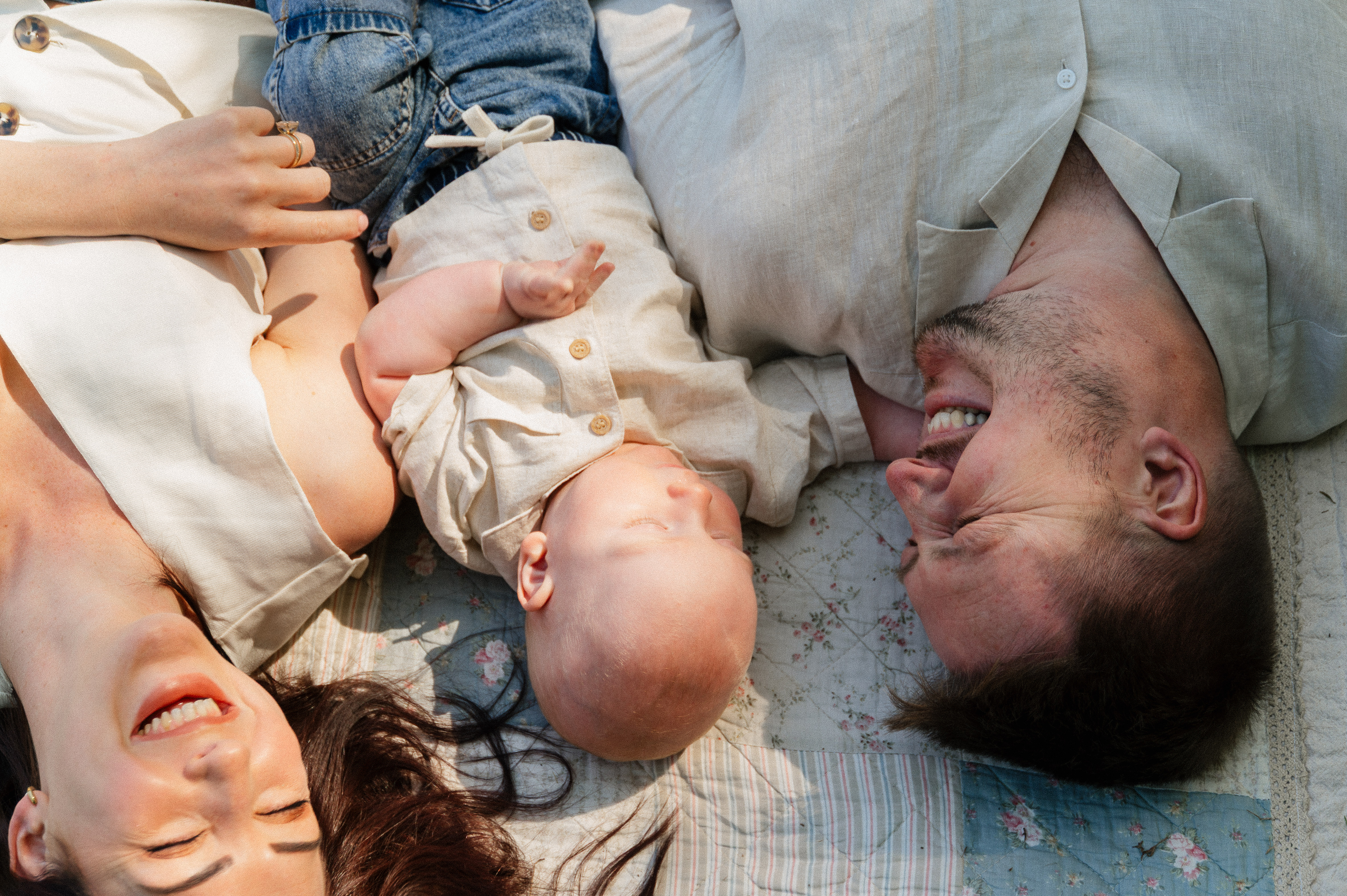 mom, dad and 6-month old son lying on a blanket outside on their backs laughing.