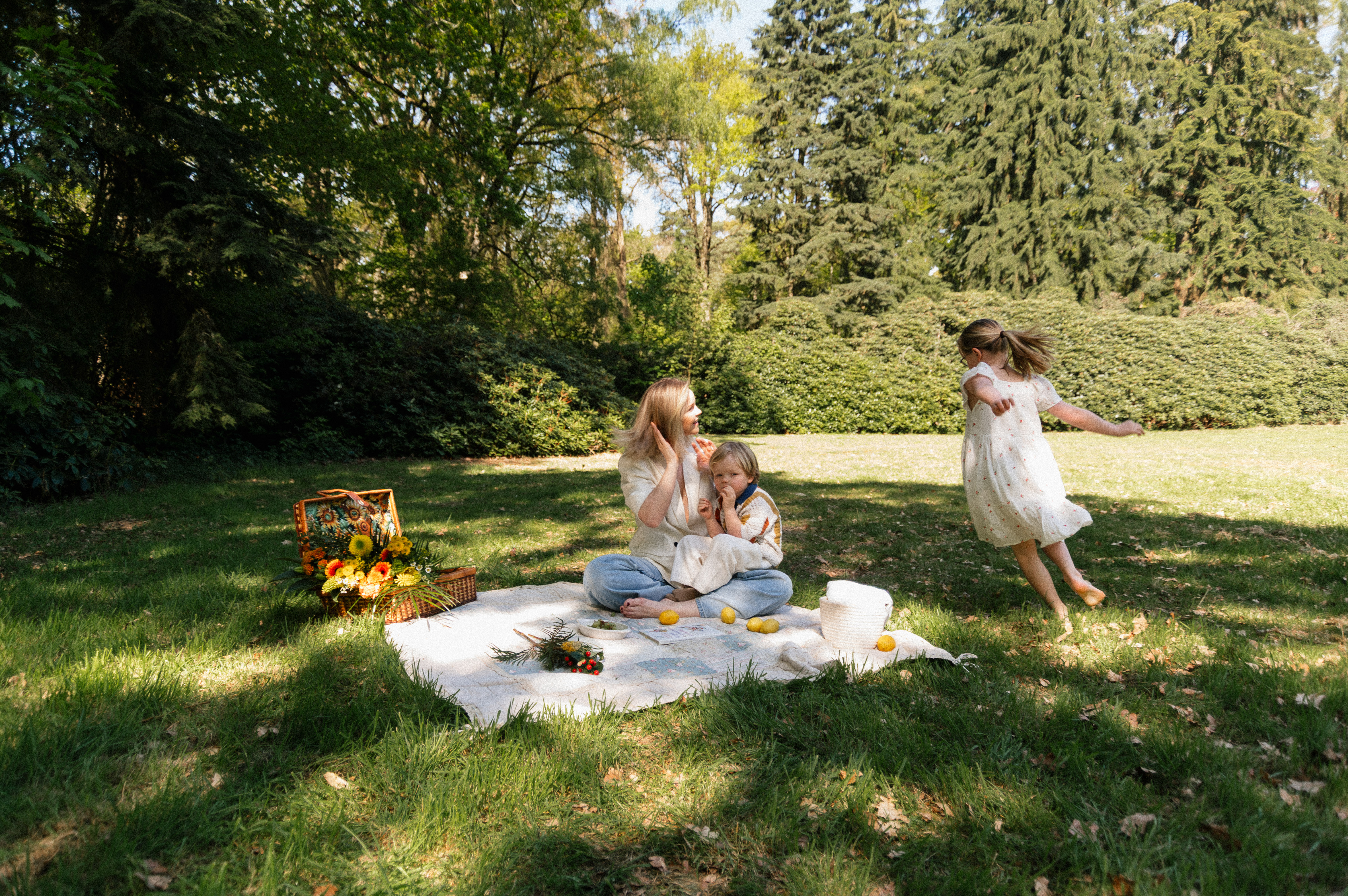 wide photo of mom sitting on picnic blanket with 3 year old son on her lap and 6 year old daughter running around them.