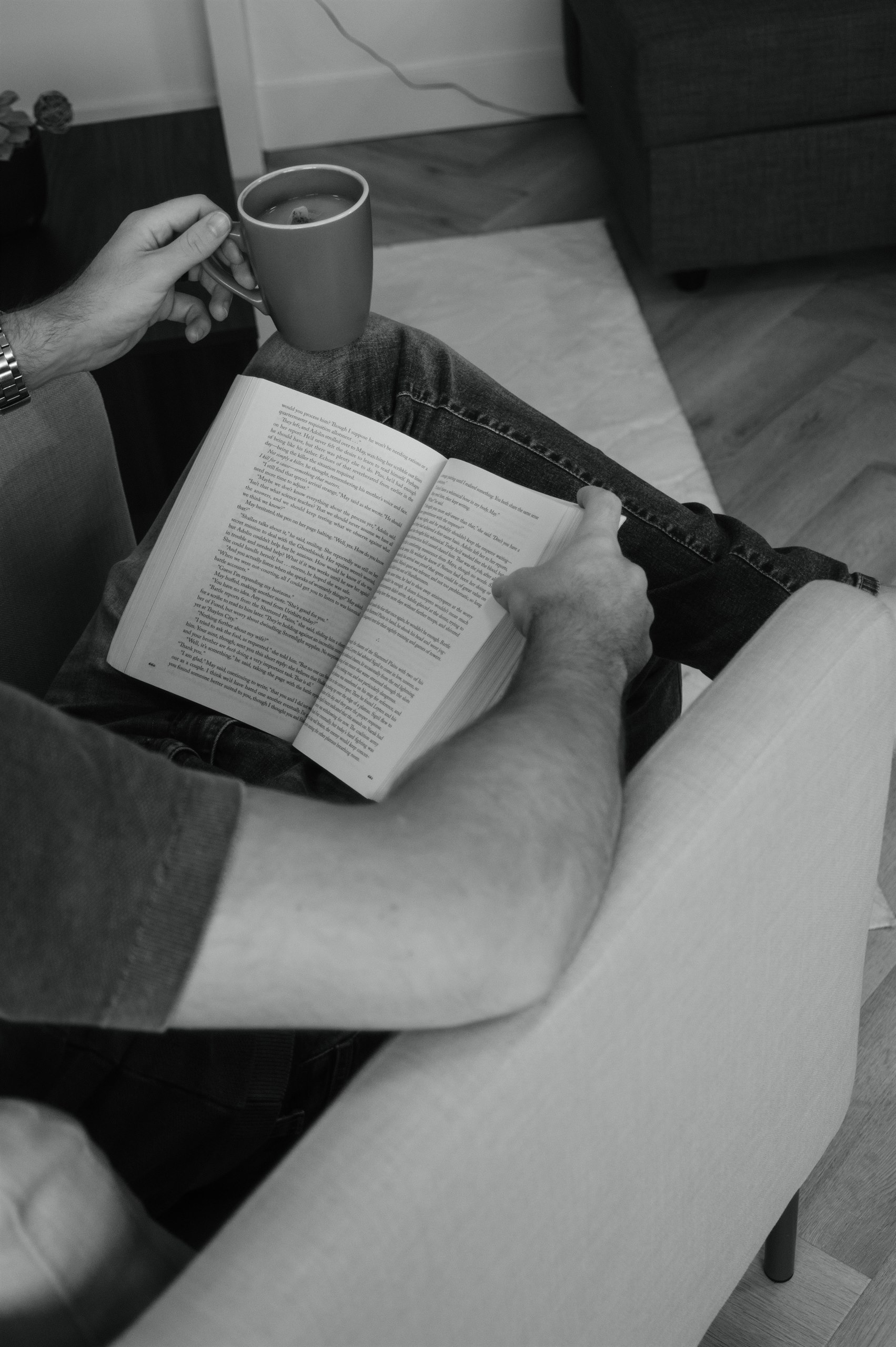 Black-and-white detail of someone reading a book with a coffee nearby at home.