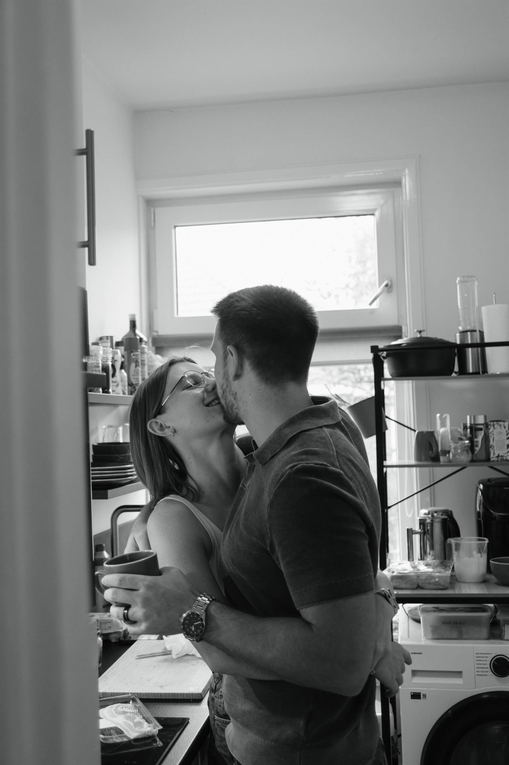 Black-and-white candid of a couple embracing in their kitchen.
