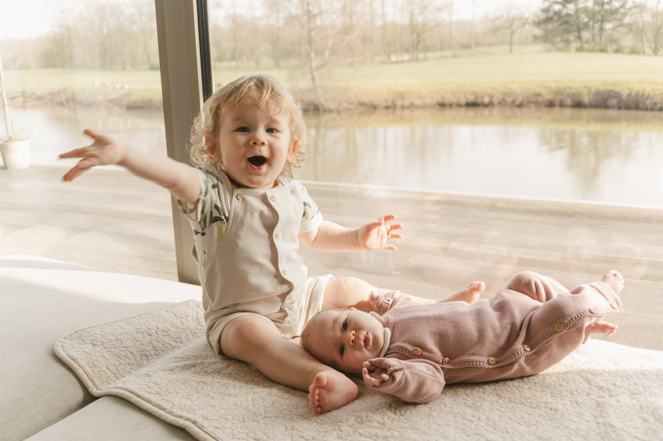 Toddler sitting beside a newborn on a blanket by a bright window.