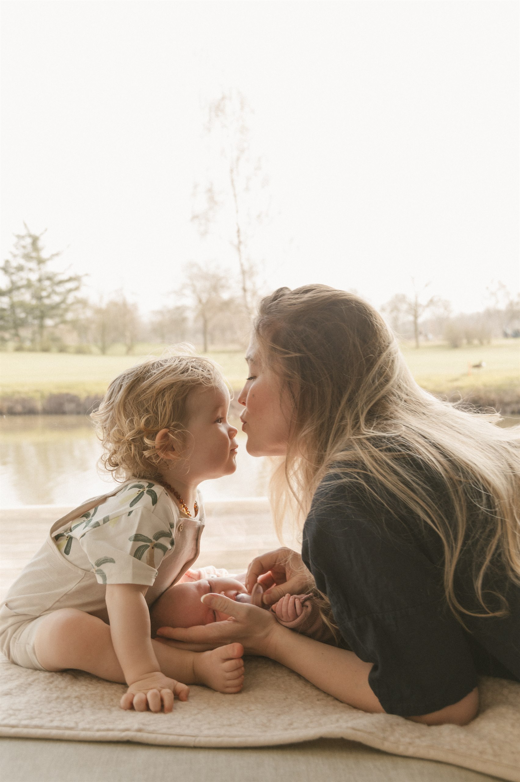 Mother leaning in to kiss her toddler while the baby lies beside them.