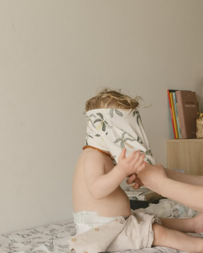 Toddler sitting on a bed with a shirt pulled over their head.