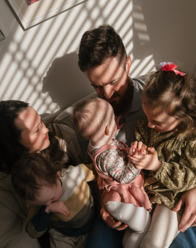 Parents and children gathered around a baby in striped window light.