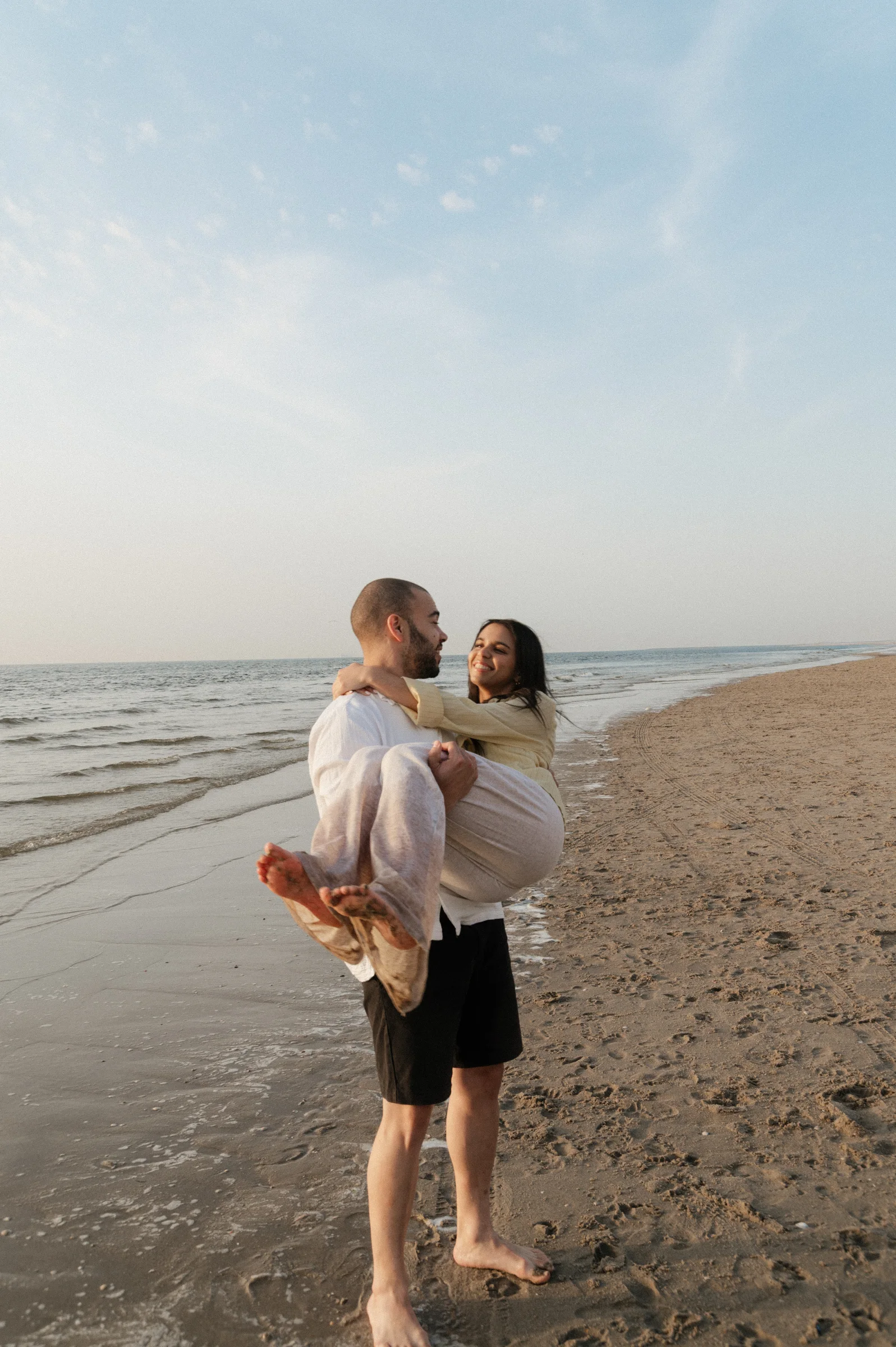 Boyfriend picking up girlfriend on the beach, looking at each other with the ocean in the background.