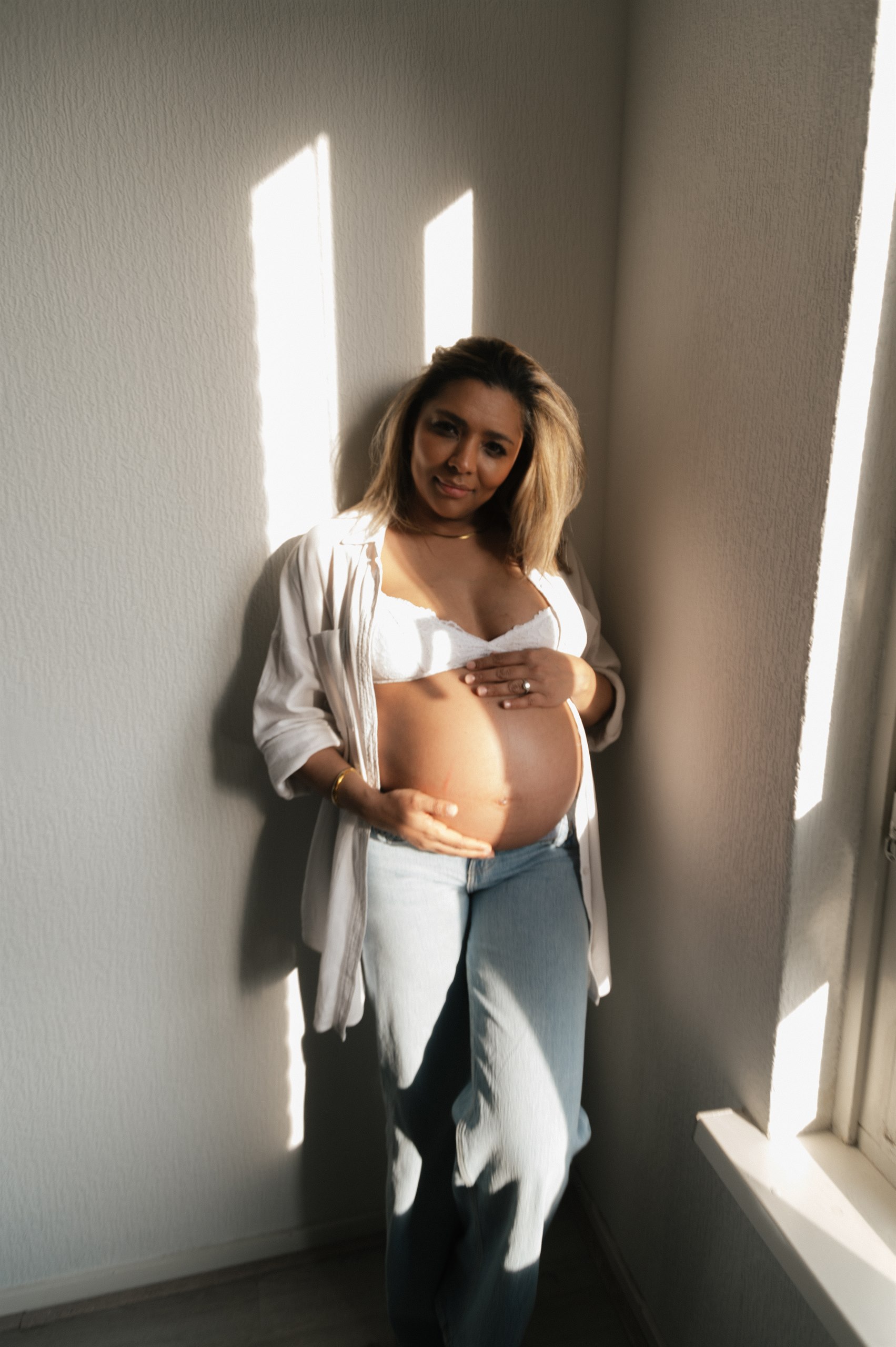 Expectant mother standing by a window with sunlight across the wall.