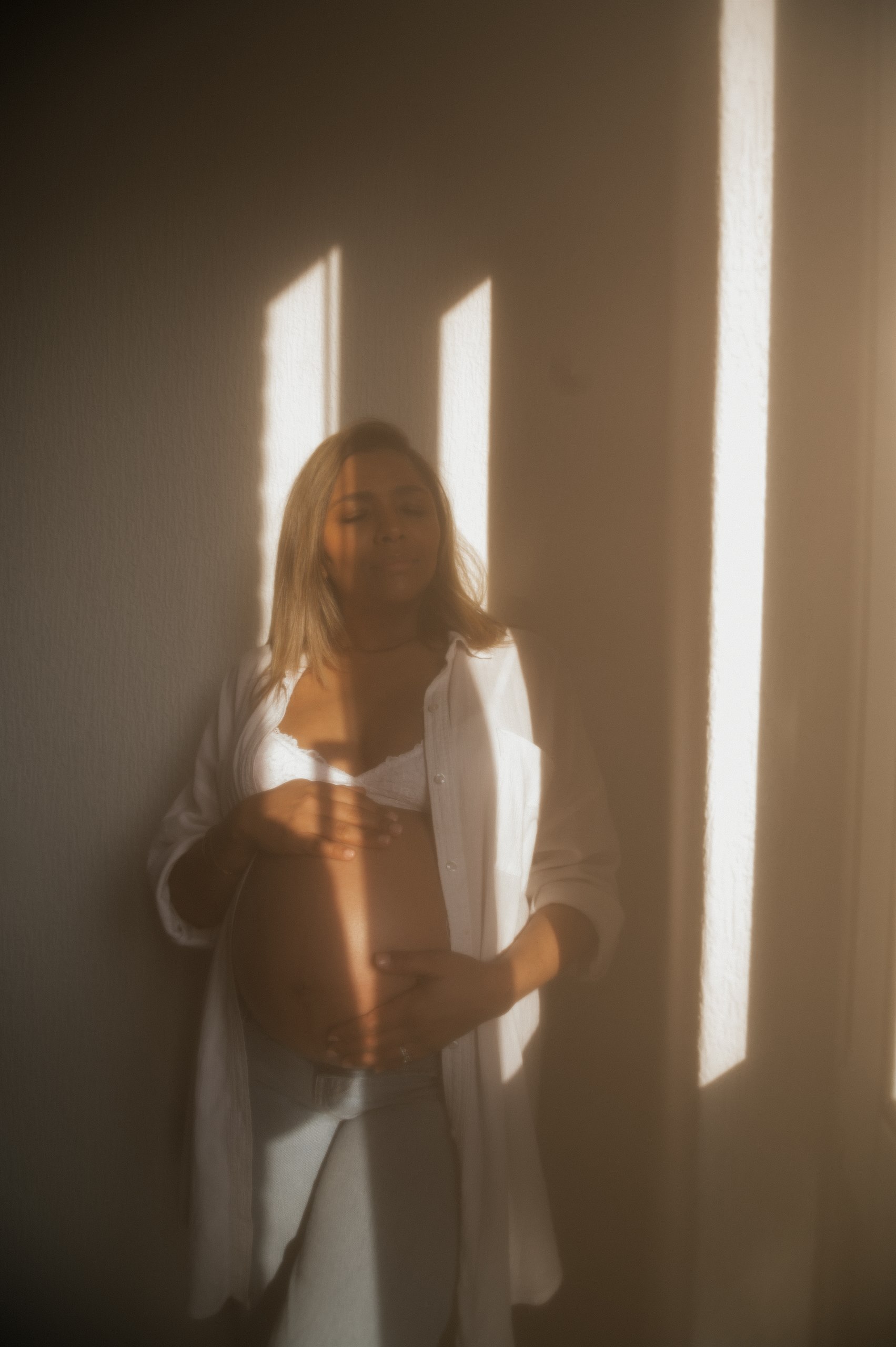 Expectant mother standing in striped window light with her hands on her belly.