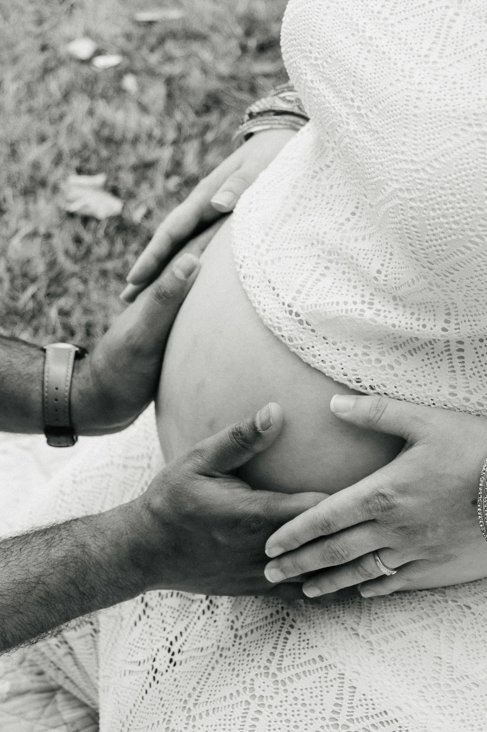 black & white photo of pregnant mom sitting with her hands and dads hand on her belly